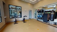 This fitness center is thoughtfully designed to accommodate a wide range of workout styles across its single-floor layout. The space is divided into distinct functional zones, each offering a specialized training environment. The main cardio area features Matrix treadmills and elliptical machines positioned along expansive floor-to-ceiling windows that frame views of palm trees and the surrounding landscape, flooding the room with natural light and creating an open, airy atmosphere. Soaring ceilings with clerestory windows further enhance the sense of volume, while pendant lighting and recessed fixtures provide ample illumination throughout.

A dedicated functional training studio houses spin bikes, TRX suspension trainers, battle ropes, and a well-stocked equipment rack featuring kettlebells, medicine balls, and stability balls. Large wall-mounted mirrors and a framed window into the adjacent space contribute to a visually expansive feel. The studio floor is finished with durable speckled rubberized flooring, while the cardio and strength areas feature warm hardwood-style flooring.

The strength training zone is generously outfitted with cable machines, selectorized weight stations, and free weight equipment, all arranged to maximize usability. A decorative geometric tile accent wall adds visual interest, complementing the clean, neutral tones of the overall design. The center also includes a separate entry point and restroom access, ensuring convenient, self-contained use.