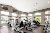 This fitness center offers a spacious, single-floor layout designed to support a full range of workout routines. The room features soaring vaulted ceilings with recessed lighting and multiple ceiling fans, creating a bright and well-ventilated environment. Large arched windows wrap around the perimeter, flooding the interior with natural light and offering views of the surrounding outdoor greenery and pool area.

The flooring throughout is rich hardwood, providing a clean, durable surface beneath the equipment. A striking herringbone-patterned wood accent wall serves as a notable design focal point, adding a warm, contemporary touch to the space.

Cardio equipment is well-represented, with multiple treadmills, elliptical trainers, a StairMaster, and stationary bikes arranged across the floor on protective rubber mats. Strength training options include a multi-station cable machine and a rack of free weights, accommodating both resistance and functional training needs. Wall-mounted mirrors along one side visually expand the room and allow users to monitor their form.

Mounted televisions provide entertainment options during workouts, while the abundance of windows ensures the space remains airy and energizing. Overall, the fitness center combines practical functionality with thoughtful design elements for a comfortable workout experience.