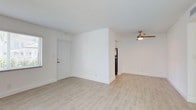 This single-floor residence presents a clean, minimalist aesthetic defined by a consistent palette of crisp white walls, white trim, and light wood-look tile flooring that runs throughout the entire unit. The open living area is generously proportioned, benefiting from natural light admitted through blinded windows near the front entry door. A ceiling fan with integrated lighting anchors the main living space, providing both comfort and illumination. A textured ceiling adds subtle visual interest overhead.

The kitchen is positioned adjacent to the living area and is partially separated by a structural half-wall, creating an open yet defined layout. It features white cabinetry, granite-look countertops, a stainless steel range, and a sink positioned beneath a window — allowing natural light into the workspace. Track lighting above the kitchen provides focused task illumination.

The bedroom areas are well-proportioned, each with the same light flooring and bright white walls. One bedroom includes a closet with a rod and open shelving, while another features a window with a view of exterior greenery, filling the room with natural light. Doorways connect rooms efficiently, and a bathroom is accessible from the bedroom corridor. The overall ambiance is clean, bright, and move-in ready.
