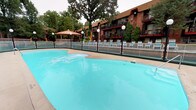 This outdoor swimming pool area is a well-appointed recreational facility set within the courtyard of a multi-story brick building. The centerpiece is a generously sized freeform pool with clear, light blue water, accessible via built-in steps and surrounded by a concrete pool deck enclosed within a chain-link safety fence. Globe-topped light poles are positioned throughout the perimeter, providing illumination for evening use.

A wooden sundeck borders one side of the pool area, furnished with rows of lounge chairs ideal for relaxing. A covered gazebo with a conical roof serves as a distinctive focal point within the complex's landscaped grounds. Notable decorative elements include a carved peacock sculpture and a mermaid statue, adding character to the space.

A brick poolside bar structure features an interior with terracotta tile flooring, a wide service counter with bar seating, and large windows overlooking the pool — making it a functional social hub. Supporting infrastructure includes a dedicated utility room housing pool mechanical and filtration equipment, as well as a supply storage room stocked with maintenance materials. Mature trees and manicured greenery throughout the grounds enhance the overall ambiance.