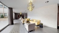 This thoughtfully designed lobby presents a welcoming and contemporary atmosphere, combining clean architectural lines with carefully selected furnishings. The space is elongated in layout, allowing for distinct seating arrangements along its length while maintaining an open, airy feel throughout.

A standout feature is the full-height textured white brick accent wall, which runs along one side of the lobby and adds depth and visual interest to the interior. Complementing this is a series of sculptural spiral chandeliers suspended from the ceiling, finished in gold tones, that serve as striking focal points and provide warm ambient lighting across the space.

Large floor-to-ceiling windows along the front of the lobby flood the interior with natural light and offer clear views of the surrounding outdoor area, creating a strong visual connection between the interior and exterior. Sheer draping curtains frame the glazing and soften the brightness when needed.

The flooring consists of light grey wood-look tiles, accented near the entrance by a geometric patterned area rug. Seating is arranged in casual groupings and includes soft yellow leather sofas, patterned armchairs, and neutral upholstered chairs, paired with small white accent tables. Potted plants are placed throughout, adding a natural touch. A staircase with a metal railing is visible toward the rear of the space.