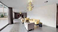 This thoughtfully designed lobby presents a welcoming and contemporary atmosphere, combining clean architectural lines with carefully selected furnishings. The space is elongated in layout, allowing for distinct seating arrangements along its length while maintaining an open, airy feel throughout.

A standout feature is the full-height textured white brick accent wall, which runs along one side of the lobby and adds depth and visual interest to the interior. Complementing this is a series of sculptural spiral chandeliers suspended from the ceiling, finished in gold tones, that serve as striking focal points and provide warm ambient lighting across the space.

Large floor-to-ceiling windows along the front of the lobby flood the interior with natural light and offer clear views of the surrounding outdoor area, creating a strong visual connection between the interior and exterior. Sheer draping curtains frame the glazing and soften the brightness when needed.

The flooring consists of light grey wood-look tiles, accented near the entrance by a geometric patterned area rug. Seating is arranged in casual groupings and includes soft yellow leather sofas, patterned armchairs, and neutral upholstered chairs, paired with small white accent tables. Potted plants are placed throughout, adding a natural touch. A staircase with a metal railing is visible toward the rear of the space.