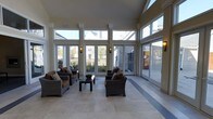 This single-floor space is a beautifully appointed sunroom lounge designed to maximize natural light and a strong connection to the outdoors. Expansive floor-to-ceiling windows and transom glazing line the walls, flooding the interior with daylight and offering sweeping views of the surrounding landscaped grounds and neighborhood beyond. The generous ceiling height amplifies the sense of openness, while decorative pilasters and crown molding details lend architectural refinement to the space.

The flooring consists of large-format light tile laid in a clean grid pattern, with a contrasting darker inset border that subtly defines the central seating area. Furnishings include multiple wicker lounge chairs with warm brown cushions and patterned throw pillows, arranged around a low wicker coffee table — creating an inviting and comfortable gathering environment.

Wall-mounted sconce lighting complements the abundant natural light, ensuring the space remains well-lit at any time of day. Multiple French doors and sliding glass doors provide direct access to the exterior, enhancing ventilation and ease of flow between indoors and outdoors. The overall ambiance is relaxed yet polished, making this lounge well-suited for casual socializing or quiet relaxation.