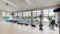 Located at 3280 Mill Run Dr in Hilliard, Ohio, this fitness center is a thoughtfully designed, single-floor facility built to support a comprehensive workout experience. The space is bathed in natural light, courtesy of expansive floor-to-ceiling windows that line the walls and offer views of the surrounding residential community, complete with lush greenery and a pool area visible in the background.

The cardio zone is prominently featured along the windowed wall, housing a row of treadmills, elliptical trainers, and stationary bikes — all equipped with individual display screens for entertainment during workouts. A dedicated strength training area offers a robust cable and pulley machine system, a full rack of dumbbells in varying weights, and multiple selectorized resistance machines suited for targeted muscle training.

The interior design features warm, neutral-toned walls paired with light, durable flooring that gives the space a clean and open feel. Vaulted ceilings with recessed lighting and ceiling fans enhance the airy ambiance. Full-length mirrors strategically placed along one wall amplify the sense of space while providing visual feedback during exercise. The overall layout is spacious and well-organized, allowing for comfortable movement between zones and accommodating multiple users simultaneously.