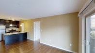 This single-floor residential space offers a clean, functional layout designed for comfortable everyday living. The main living area is finished with warm hardwood-style flooring that runs throughout, providing a cohesive and inviting foundation for the space. Smooth white ceilings contrast neatly with the warm beige-toned walls, creating a bright and airy ambiance.

A partial view into the kitchen area reveals a dark-topped counter or breakfast bar, complemented by pendant lighting overhead, suggesting a practical and stylish cooking and dining space that connects openly to the main living area. This open-concept flow between the kitchen and living space enhances the sense of roominess and encourages natural movement throughout.

A white interior doorway visible from the living area provides access to an adjacent room, hinting at a thoughtful and organized floor plan. Clean white baseboards trim the walls throughout, adding a crisp, finished look to the interior.

The space is well-equipped with multiple electrical outlets and a ceiling vent, reflecting practical, move-in-ready functionality. Overall, the interior presents a neutral, versatile palette that serves as an excellent canvas for a variety of interior design styles.