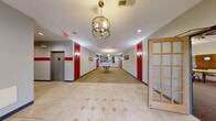 This veteran-focused nonprofit community facility offers a well-appointed, single-floor layout designed to serve and welcome its members. The main corridor makes a strong first impression with large-format tile flooring accented by a geometric mosaic pattern, bold red wall panels, and warm wall sconces flanking the hallway. An elegant brass orb pendant light hangs overhead, adding a refined touch to the entryway, while an American flag and a curated photo display at the corridor's end reinforce the space's patriotic identity.

The lobby area features carpeted flooring, pendant lighting, and a central round table stocked with informational materials, serving as a welcoming point of contact for visitors. Elevator access and glass-paned French doors provide convenient connectivity throughout.

The centerpiece of the facility is a large, open event hall with plush carpeting, multiple ceiling fans, skylights, and both recessed and pendant lighting. Rows of round tables and chairs make it well-suited for community gatherings, meetings, and events. Large windows along one wall allow natural light to filter in, creating an inviting atmosphere.

Additional spaces include a lounge area with a large-screen television and comfortable seating, a private office or meeting room with a skylight, small conference table, and office equipment, and a utility corridor with tile flooring and emergency exit access. Together, these areas form a functional and community-oriented environment.