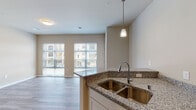 This single-floor unit offers a well-designed open-concept layout where the kitchen flows seamlessly into the living area, creating a cohesive and functional living space. The kitchen is a standout feature, equipped with white shaker-style cabinetry, speckled granite countertops, and a full suite of stainless steel appliances including a range, microwave, and dishwasher. A peninsula with a double undermount sink and pendant lighting above serves as both a workspace and a natural divider between the kitchen and living areas. Wood-style plank flooring runs throughout the unit, providing a warm, consistent aesthetic. The living area benefits from generous natural light courtesy of two large windows fitted with horizontal blinds, offering views of the surrounding neighborhood. The bathroom continues the unit's design language with matching granite countertops, white cabinetry, and wood-style flooring, complemented by a well-lit vanity mirror. Neutral wall tones throughout contribute to a clean, modern ambiance, while the open floor plan enhances the overall sense of space and connectivity between rooms.