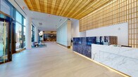 This elegant lobby presents a refined first impression through a seamless blend of premium materials and contemporary design. The reception area commands immediate attention with its dramatic desk, clad in deep blue veined marble contrasted by a crisp white marble countertop, all set beneath a signature warm-toned slatted wood ceiling that extends across the space and doubles as a textured wall feature. Polished large-format stone flooring reflects the ambient and recessed lighting, enhancing the sense of openness and grandeur throughout.

Flanking the main entrance, floor-to-ceiling glazing floods the lobby with natural light and frames views of a landscaped outdoor courtyard. Curated potted trees and planters bring a touch of greenery indoors near the entrance, while stainless steel elevator doors framed by matching marble panels provide a polished transition point within the space.

Beyond the reception, a wide corridor leads to a sophisticated lounge area featuring sculptural globe pendant lighting, soft seating arrangements in muted tones, and bold teal accents in rugs and upholstery. Glass-partitioned meeting nooks and a feature brick accent wall add depth and functionality. Artwork lines the gallery-style hallway walls, reinforcing the space's refined, professional character throughout.