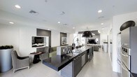 This contemporary single-floor space showcases a thoughtfully designed open-plan layout that seamlessly blends kitchen, dining, and living areas into one cohesive environment. The kitchen serves as the focal point, anchored by a substantial dark granite island with an undermount sink, dark cabinetry, and a bold pendant light fixture that adds a sculptural element to the space. The island is complemented by stylish bar seating, making it ideal for casual dining or entertaining. The living area features built-in niche displays set against a textured white accent wall, providing visual depth and an artistic touch. Comfortable seating arrangements are thoughtfully positioned throughout, creating distinct yet connected zones. A large flat-screen television is integrated into the open layout, enhancing the space's functionality. Large-format light-toned floor tiles run throughout, reflecting the abundant recessed lighting overhead and contributing to the bright, airy ambiance. The overall design palette of crisp whites, dark browns, and neutral tones gives the space a polished, upscale character, while the open-concept configuration maximizes the sense of flow and spaciousness throughout.