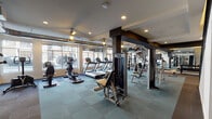 Located at 424 Benson Ave NE in Grand Rapids, Michigan, this two-story fitness facility is thoughtfully designed to accommodate a wide range of workout styles and training needs. The space is organized across two levels connected by a staircase, with each floor dedicated to distinct fitness activities.

The main floor houses a well-equipped cardio zone featuring multiple treadmills, ellipticals, and stationary bikes positioned along large windows that allow generous natural light and views of the surrounding neighborhood. Mirrored walls amplify the sense of space throughout this level. A strength training area includes weight benches, a barbell rack with plate weights, a cable machine, and a dumbbell rack, all set on durable, patterned floor tiles. Exposed ductwork adds an industrial aesthetic to portions of the space.

The upper level features a dedicated group exercise and studio area with hardwood-style flooring, ceiling fans, and expansive windows. This room is stocked with spin bikes, resistance bars, exercise balls, TRX bands, and stacked step platforms, making it ideal for group classes or functional training sessions. A separate utility area with warm-toned wood cabinetry, open shelving, and a dark countertop provides a practical support space within the facility.