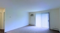 This single-floor space offers a clean and functional layout designed for comfortable everyday living. The main living area is finished with wall-to-wall carpeting that adds warmth underfoot and contributes to a quiet, cozy atmosphere throughout. Neutral white walls keep the space feeling bright and open, providing a versatile backdrop for a variety of furnishing styles.

A prominent sliding glass door dominates one wall of the living area, fitted with full-length vertical blinds that allow for adjustable light control and privacy. When open, the blinds allow natural light to fill the room generously. A wall-mounted air conditioning unit is installed nearby, ensuring climate comfort during warmer months, while a baseboard heating unit along the lower wall provides warmth during cooler seasons. Together, these features offer year-round temperature control.

The overall ambiance of the space is simple, functional, and move-in ready, with a neutral palette that makes it easy to personalize. The single-floor layout ensures seamless accessibility and ease of navigation throughout. This space is well-suited for those seeking a practical, low-maintenance living environment with essential amenities already in place.