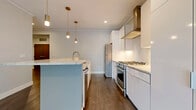 Located at 19 North St in Buffalo, NY, this single-floor residence delivers a refined modern aesthetic with high-quality finishes throughout. The chef's kitchen is a standout centerpiece, showcasing floor-to-ceiling white gloss cabinetry, a stainless steel gas range with a professional hood, a stainless steel refrigerator, and a generous island topped with granite countertops. Brass pendant lights add a warm, sophisticated accent overhead, while dark hardwood flooring grounds the space with richness and contrast. The open layout flows naturally from the kitchen into a spacious living area bathed in natural light through large, black-framed floor-to-ceiling windows that frame views of the surrounding neighborhood's mature trees. Additional rooms feature plush patterned carpeting and black-framed windows that carry the design language throughout. The bathroom is elegantly appointed with large-format marble-look tile wrapping a glass-enclosed walk-in shower, complemented by hexagonal tile flooring and dark wood vanity cabinetry with brass hardware accents. Neutral wall tones and clean white trim are consistent throughout, creating a cohesive, contemporary atmosphere. Recessed lighting provides even illumination across all living areas, completing this well-appointed and thoughtfully designed urban residence.