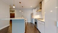 Located at 19 North St in Buffalo, NY, this single-floor residence delivers a refined modern aesthetic with high-quality finishes throughout. The chef's kitchen is a standout centerpiece, showcasing floor-to-ceiling white gloss cabinetry, a stainless steel gas range with a professional hood, a stainless steel refrigerator, and a generous island topped with granite countertops. Brass pendant lights add a warm, sophisticated accent overhead, while dark hardwood flooring grounds the space with richness and contrast. The open layout flows naturally from the kitchen into a spacious living area bathed in natural light through large, black-framed floor-to-ceiling windows that frame views of the surrounding neighborhood's mature trees. Additional rooms feature plush patterned carpeting and black-framed windows that carry the design language throughout. The bathroom is elegantly appointed with large-format marble-look tile wrapping a glass-enclosed walk-in shower, complemented by hexagonal tile flooring and dark wood vanity cabinetry with brass hardware accents. Neutral wall tones and clean white trim are consistent throughout, creating a cohesive, contemporary atmosphere. Recessed lighting provides even illumination across all living areas, completing this well-appointed and thoughtfully designed urban residence.