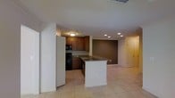 This single-floor residence offers a thoughtfully designed open-concept layout that seamlessly connects the kitchen and living areas, creating a spacious and functional environment. The kitchen is a standout feature, showcasing rich medium-toned wood cabinetry that extends to the upper walls, complemented by classic subway tile backsplash and granite countertops. A center island with a white base and granite surface serves as both a prep space and a casual gathering point, enhancing the kitchen's practicality and visual appeal. Black appliances, including a built-in oven and microwave, add a sleek, cohesive touch to the culinary space. The flooring throughout is finished in light-toned ceramic tile, providing a clean and durable surface that ties the open areas together. Recessed lighting in the living area, paired with a flush-mount ceiling fixture in the kitchen, ensures the space is well-lit throughout. A deep, earthy brown accent wall anchors the transition between the kitchen and the adjacent living space, adding a warm, grounding contrast to the otherwise neutral palette. Doorways leading to additional areas of the home are visible, suggesting a well-connected and practical floor plan designed for comfortable everyday living.
