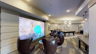 The game room is a well-appointed, single-floor amenity space designed to accommodate a variety of recreational activities. The room is anchored by a striking large-format video wall composed of multiple display panels, making it an ideal focal point for gaming, streaming, or social viewing. In front of the video wall, a cluster of curved, high-back leather lounge chairs arranged around a low ottoman creates an inviting and stylish seating area.

The space transitions seamlessly toward a dedicated billiards area, where a full-size pool table takes center stage beneath a pair of geometric pendant light fixtures that add a modern, upscale touch. The billiards zone is further complemented by framed wall art and decorative accents that contribute to the room's polished aesthetic.

Recessed ceiling lighting throughout provides consistent, even illumination, while a tray ceiling detail adds architectural depth. The flooring features a patterned carpet with linear accents, grounding the space with a contemporary design. The overall ambiance is sophisticated yet relaxed, balancing a lounge-like atmosphere with practical entertainment functionality.