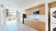 Located at 2701 Shattuck Ave in Berkeley, California, this thoughtfully designed single-floor unit offers an efficient and modern living environment. Light wood-look flooring runs throughout, complementing the clean, neutral palette of white walls and crisp finishes that give the space an airy, contemporary feel.

The open-plan kitchen and living area forms the heart of the unit. The kitchen is outfitted with warm honey-toned wood cabinetry — both upper and lower — paired with light countertops, a built-in cooktop, an over-the-range microwave, and a full-size refrigerator. A stainless steel finish is available on select appliances, adding a modern touch. A dedicated nook houses a stacked in-unit washer and dryer, providing added convenience.

The living area benefits from expansive windows that flood the space with natural light and frame pleasant views of the surrounding Berkeley neighborhood, including lush treetops and the scenic East Bay hills. A wall-mounted mini-split HVAC system ensures year-round climate comfort.

The bathroom is clean and functional, featuring a soaking tub with a tiled surround, a toilet, and a wall-mounted sink. Accessibility features such as grab bars are also present, reflecting inclusive design. Light wood-look flooring continues here, maintaining a cohesive aesthetic throughout the unit.