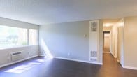 This single-floor space presents a clean, functional layout with notable structural and design details throughout. The main living area features dark hardwood flooring that contrasts with light gray walls, creating a balanced, neutral aesthetic. A textured popcorn ceiling adds a classic touch overhead. Natural light filters in through a window on the left wall, where sheer curtains allow soft, diffused sunlight to brighten the room. A built-in wall-mounted HVAC unit with upper and lower vents is integrated directly into the wall, providing a space-efficient heating and cooling solution. White baseboards trim the perimeter of the room, offering a clean delineation between the walls and flooring. An open doorway on the right side of the main room leads to an adjacent space, suggesting a connected, flowing floor plan. The overall ambiance is understated and move-in ready, with a neutral palette that provides a versatile backdrop adaptable to a variety of interior styles. The space offers a straightforward, practical design suited for comfortable everyday living.