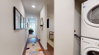 This well-appointed single-floor residence offers a thoughtfully designed layout that balances functionality with modern style. A welcoming hallway, accented by a colorful geometric runner rug and a gold-framed mirror, leads into the heart of the home. The kitchen is equipped with stainless steel appliances, including a range, microwave, and dishwasher, and features a two-tone cabinet design pairing crisp white upper cabinets with dark espresso lower cabinetry. A sleek tile backsplash and light countertops complete the polished look, while wood-style flooring flows seamlessly into the adjoining dining and living areas. The living room is bright and airy, benefiting from a wall-mounted mini-split system and a sliding glass door that opens to a private balcony, allowing ample natural light to fill the space. The bedroom is a cozy retreat with neutral tones, a wall-mounted television, and direct access to the corridor. The full bathroom is finished with large-format tile surrounding a soaking tub and shower combination, a dark-cabinetry vanity with a white countertop, and a generously sized mirror. Throughout the residence, recessed lighting, clean white walls, and wood-style flooring create a cohesive, contemporary ambiance.