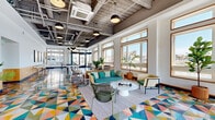Located at Harbor Club in Baltimore's vibrant 21231 waterfront district, this single-floor amenity space offers a generously sized, open-plan layout designed for socializing and recreation. The most striking design element is the bold, multicolored geometric tile flooring—a kaleidoscope of triangular shapes in teal, coral, yellow, and white—that anchors the entire space visually.

Soaring exposed ceilings reveal industrial-style ductwork and concrete beams, contrasted by sleek pendant lighting suspended at varying heights. Large wood-framed windows line the perimeter, flooding the interior with abundant natural light and framing elevated views of Baltimore's cityscape and harbor marina.

The space is organized into distinct functional zones. A lounge area features teal upholstered sofas, woven accent chairs, and a round white coffee table arranged on a patterned area rug. An adjacent games zone includes a full-size pool table and shuffleboard. A bar and dining area offers a marble-topped counter, woven barsack seating, wood cabinetry, and a wall-mounted television. A glass-enclosed corner room provides direct access to an outdoor terrace with marina views. Potted tropical plants throughout add warmth, completing the lively, modern ambiance.