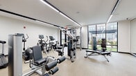 Located at 308 NJ-31 N in Flemington, New Jersey, this contemporary fitness center offers a thoughtfully designed, single-level workout facility. The space is bathed in natural light, courtesy of expansive floor-to-ceiling windows framed in sleek black steel, which provide views of the surrounding outdoor amenity areas and lend an open, airy ambiance to the interior.

Linear LED lighting fixtures run across the white ceiling, ensuring consistent, bright illumination throughout the space. The flooring features a durable, speckled rubberized surface suited for high-traffic fitness use.

The facility is comprehensively equipped for a full-body workout. Cardio options include Life Fitness treadmills and elliptical cross-trainers, each outfitted with digital display consoles. Strength training is well-represented with a multi-station cable machine, a cable pull system, a leg extension and curl machine, a functional trainer, and a rowing machine. A dedicated free weights area includes a full dumbbell rack and an adjustable workout bench, complemented by a large wall mirror and a mounted flat-screen television.

Glass-paneled partitions and doors connect to an adjacent room visible from the interior, adding a sense of depth to the layout. The overall design is clean, modern, and functional, creating a professional fitness environment.