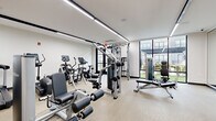 Located at 308 NJ-31 N in Flemington, New Jersey, this contemporary fitness center offers a thoughtfully designed, single-level workout facility. The space is bathed in natural light, courtesy of expansive floor-to-ceiling windows framed in sleek black steel, which provide views of the surrounding outdoor amenity areas and lend an open, airy ambiance to the interior.

Linear LED lighting fixtures run across the white ceiling, ensuring consistent, bright illumination throughout the space. The flooring features a durable, speckled rubberized surface suited for high-traffic fitness use.

The facility is comprehensively equipped for a full-body workout. Cardio options include Life Fitness treadmills and elliptical cross-trainers, each outfitted with digital display consoles. Strength training is well-represented with a multi-station cable machine, a cable pull system, a leg extension and curl machine, a functional trainer, and a rowing machine. A dedicated free weights area includes a full dumbbell rack and an adjustable workout bench, complemented by a large wall mirror and a mounted flat-screen television.

Glass-paneled partitions and doors connect to an adjacent room visible from the interior, adding a sense of depth to the layout. The overall design is clean, modern, and functional, creating a professional fitness environment.