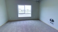 This single-floor space offers a clean, straightforward layout designed for comfortable everyday living. The interior is finished with wall-to-wall carpeting in a neutral tone, providing a soft and inviting underfoot feel throughout the space. Walls are painted in a calm, muted gray-green hue that contributes to a quiet and understated ambiance.

A standout feature is the large sliding window that fills the room with natural light while framing views of the surrounding outdoor environment, including neighboring buildings, open lawn areas, and a small body of water. This connection to the outside enhances the sense of openness within the space.

Subtle architectural details include a ceiling-mounted track or industrial-style rail element visible near the ceiling, adding a minor utilitarian design note. The baseboard trim provides a clean transition between the walls and the carpeted floor. Overall, the space presents a simple, functional interior with neutral finishes, natural light, and pleasant outdoor views — making it a versatile and practical environment.