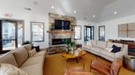 This single-floor communal space is designed with a clean, modern aesthetic that balances comfort and functionality. Polished concrete floors run throughout, reflecting natural light that pours in through large, gray-trimmed windows fitted with plantation blinds.

The central lounge area serves as the heart of the space, anchored by a dramatic floor-to-ceiling stacked stone fireplace with a rustic wood mantel and wall-mounted television. Comfortable neutral sofas and leather accent chairs are arranged around a warm-toned area rug, creating an inviting atmosphere for relaxation or socializing.

Adjacent to the lounge, a fully equipped kitchen features a large island with dark granite countertops and leather-upholstered bar seating, white shaker cabinetry, shiplap accent walls, stainless steel appliances including a refrigerator and microwave, and ample counter space. Glass-paneled sliding doors connect the kitchen to the broader open-plan layout, enhancing the sense of flow.

A dedicated dining nook provides a bright, sunlit setting with a round table and metal chairs, framed by contemporary artwork and tall indoor plants. Additional seating near the exterior glass doors allows for quiet, light-filled moments. The space opens directly to an outdoor area, reinforcing its open and connected design throughout.