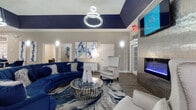 This single-floor space serves as a beautifully appointed clubhouse or leasing office, designed with a cohesive and contemporary aesthetic that blends bold color with refined finishes. The interior is defined by a striking navy blue and neutral gray color palette carried throughout, from the tray ceilings accented in deep navy to the plush velvet furnishings and abstract artwork adorning the walls.

The main lounge area is a standout feature, centered around a large curved navy sectional sofa paired with a mirrored, cylindrical coffee table atop a blue-and-white marbled area rug. A modern ring chandelier illuminates the seating area, while a wall-mounted electric fireplace with a metallic tile surround and an overhead flat-screen TV create a dynamic focal point. A sleek wingback accent chair completes the arrangement.

Adjacent to the lounge is a dedicated workspace or meeting area, furnished with a glass-top desk and coordinating navy velvet chairs, ideal for consultations or office use. French doors along the perimeter allow natural light to pour in and provide views of the outdoors, enhancing the open, airy feel of the space.

Wide-plank hardwood-style flooring runs continuously throughout, tying each area together seamlessly. Transom windows above interior doors add architectural interest, while carefully selected abstract art pieces reinforce the space's polished, upscale character.