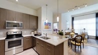 This single-floor residence showcases a thoughtfully designed open-concept layout that integrates the kitchen, dining, and living areas into one cohesive, flowing space. The kitchen is outfitted with rich, dark wood cabinetry featuring sleek hardware, complemented by a light-toned countertop that extends into a peninsula with an undermount double sink. A tile backsplash adds texture, while stainless steel appliances, including a built-in microwave and dishwasher, contribute to the space's modern functionality. Two slender pendant lights hang above the peninsula, providing focused task lighting and adding a contemporary design touch. The high ceilings amplify the sense of openness and volume throughout. Track lighting in the living area ensures flexible illumination, while large windows allow natural light to filter in, brightening the neutral color palette of cream and warm wood tones. The adjoining dining area accommodates a full table setting and transitions naturally into the living space, which is visible in the background. Light wood-style flooring runs throughout, tying the areas together with a clean, cohesive aesthetic. Colorful wall art adds personality and visual interest.