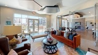 This expansive lounge is thoughtfully designed across a single level, offering a variety of distinct functional zones that cater to both relaxed socializing and more structured gatherings. The main seating area features plush, richly upholstered furniture in warm burnt orange and cool navy tones, centered around a sculptural round coffee table atop a layered area rug. Antique mirrored wall panels and angular ceiling details with geometric cutouts add architectural drama throughout.

Large floor-to-ceiling windows and glass doors flood the space with natural light and provide views of an outdoor terrace and surrounding community. The billiards zone is anchored by a sleek pool table beneath a striking ceiling installation of staggered pendant lights, set against warm wood-look tile flooring that runs throughout much of the space.

A private dining area features a long communal table, pendant lighting, and a floor-to-ceiling tiled fireplace wall with an integrated flat-screen TV, creating an intimate yet refined setting. Adjacent to this is a fully equipped wet bar with navy cabinetry, a beverage refrigerator, under-counter appliances, and a textured metallic tile accent wall. Together, these zones create a cohesive, upscale communal environment suited for a wide range of social activities.