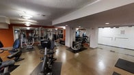 This fitness center is laid out across a single level, offering a practical and functional space for exercise. The room features light-toned flooring with rubber mats strategically placed beneath equipment to protect surfaces and ensure user safety. The textured ceiling reflects overhead lighting, providing ample illumination throughout the space. Structural columns divide the room into distinct workout zones, each outfitted with various strength training machines, including cable and weight systems. Large mirrors mounted along the accent walls, highlighted in a warm terracotta-orange tone, visually expand the space and allow users to monitor their form during workouts. Cardio equipment, including a stationary bike and an elliptical machine, is positioned toward the front of the room for easy accessibility. A glass-paneled door or partition is visible along one side, contributing to an open and airy feel. Dumbbell racks and free weight stations are also present, making this a versatile space suitable for a wide range of fitness routines. The overall ambiance is practical and focused, with a clean, organized layout that maximizes usability.