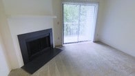 This single-story space presents a clean, functional living area designed with comfort in mind. The room's most notable feature is a sleek, dark-framed fireplace set into a white surround, providing a classic focal point that anchors the space. Neutral, light-toned carpeting spans the floor, contributing to a warm and inviting atmosphere throughout.

Natural light is a defining characteristic of this space, streaming in generously through a large sliding glass door fitted with vertical blinds. The door opens to a balcony enclosed by white railings, beyond which mature, leafy greenery creates a pleasant, private outdoor backdrop. This connection to nature enhances the overall ambiance, making the interior feel open and airy despite its modest scale.

The walls are finished in a soft, neutral tone that complements the carpet and trim, lending the space a cohesive and understated aesthetic. The simple, uncluttered design allows for flexible furniture arrangements. Overall, the combination of the fireplace, natural light, and outdoor views creates a balanced and comfortable living environment.