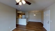 This single-floor residence offers a thoughtfully designed open-concept layout that seamlessly connects the living area and kitchen, creating a spacious and inviting atmosphere. Rich, dark hardwood flooring runs throughout the main living space, adding warmth and a classic aesthetic that anchors the interior design.

The kitchen is defined by warm wooden cabinetry, a functional peninsula with a breakfast bar, and includes standard appliances such as a refrigerator and range with a range hood above. The peninsula serves as a natural divider between the kitchen and the living area, offering both additional prep space and casual seating potential.

The living area is generously proportioned, bathed in soft ambient light from a ceiling fan with integrated lighting and a stylish pendant light that hangs gracefully in the dining zone. The textured ceiling adds subtle architectural interest overhead, while neutral-toned walls provide a versatile backdrop suitable for a variety of décor styles. A doorway at the rear of the living space suggests access to additional rooms, rounding out a functional and comfortable single-level floor plan. Overall, the space balances practical design with a warm, approachable ambiance.