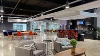 Located in the heart of Kansas City's downtown at 720 Main Street, this lower-level lounge is a generously sized, open-plan amenity space defined by its contemporary industrial aesthetic. Polished concrete floors reflect the overhead pendant and track lighting, while exposed black ceilings with visible ductwork and structural elements lend an urban, loft-like character throughout.

The layout is thoughtfully divided into distinct functional zones without sacrificing the open feel of the floor. A central lounge area features plush sofas in deep navy and rust tones, accent chairs, patterned area rugs, and rounded pouf ottomans, creating an inviting space for relaxed social interaction. Adjacent to this is a casual dining and collaboration zone with light wood tables and vibrant orange seating. A striking bar counter clad in a bold red-and-black tile mosaic anchors one side of the room, complete with bar stools and overhead pendant lighting.

Glass-walled private rooms line one perimeter, offering semi-enclosed spaces for meetings or smaller gatherings. A foosball table and a ping pong table add a recreational dimension to the space. A wall-mounted television and presentation-ready table provide functionality for group events. Elevator access is also present. The overall ambiance is energetic yet versatile, suited for both productive collaboration and social gatherings.