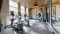 Located at 1205 Worcester St in Natick, Massachusetts, this modern fitness center offers a well-appointed, single-floor space designed for an elevated workout experience. The room is characterized by its striking warm wood-paneled ceiling, which adds a refined, contemporary aesthetic and pairs seamlessly with the neutral-toned walls and durable gray flooring throughout.

Natural light floods the space through a generous row of large windows fitted with adjustable roller shades, offering views of the surrounding outdoor landscape while maintaining a bright and energizing atmosphere. The cardio zone is well-equipped with multiple treadmills, elliptical machines, and upright bikes — each unit featuring individual display screens for an interactive workout experience.

A large floor-to-ceiling mirror along one wall visually expands the space and supports functional training, accompanied by a dedicated rack of medicine and exercise balls, yoga mats, and additional free-motion strength equipment. Recessed and linear LED lighting complements the natural light, ensuring consistent illumination throughout the day. A secured, glassed entry door provides controlled access to the facility. The overall ambiance is clean, contemporary, and purpose-built to support a comprehensive fitness routine in a polished, professional environment.