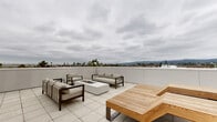 This contemporary rooftop terrace in Alameda, California offers a thoughtfully designed outdoor amenity space ideal for relaxation and entertaining. The expansive tiled deck is organized into distinct functional zones, each crafted with modern aesthetics and quality materials throughout.

At the heart of the terrace sits a sleek lounge area centered around a rectangular gas fire pit table, surrounded by neutral-toned upholstered sofas and armchairs with dark wood frames. Adjacent to this seating zone, a pair of teak slatted sun loungers provide a dedicated space for sunbathing, perfectly positioned to take in the sweeping panoramic views of the East Bay hills, surrounding neighborhoods, and distant skyline.

The outdoor kitchen area is equipped with a full-size stainless steel BBQ grill and prep station, accompanied by an outdoor television — making it well-suited for al fresco dining and gatherings. A sculptural curved teak bench adds an artistic touch near the raised Corten steel planters, which feature ornamental grasses and succulents, contributing a natural, living element to the otherwise clean, minimalist design.

Perimeter parapet walls in crisp light gray paneling frame the terrace while providing safety and a polished architectural finish. The large-format porcelain tile flooring ties the entire space together, reinforcing the modern character of this rooftop retreat.