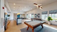 This single-floor clubroom in West Melbourne, Florida, is a thoughtfully designed communal space that blends recreational amenities with a contemporary aesthetic. The open layout is anchored by soaring vaulted ceilings that contribute to an airy, expansive atmosphere throughout. Light wood-look flooring flows across the main game area, complemented by recessed lighting and striking sculptural pendant fixtures that add a modern design element overhead.

The game zone serves as the centerpiece of the space, featuring a full-size pool table and a shuffleboard table positioned near large windows and glass doors that open to an outdoor balcony area, offering views of the surrounding landscape and palm trees. Contemporary artwork with teal and geometric motifs adorns the walls, tying together the room's coastal-modern color palette.

A well-appointed kitchen and bar area runs along one side of the space, showcasing white cabinetry, stainless steel appliances, a wall-mounted television, a vibrant blue penny-tile backsplash, and a long island with bold blue base detailing and bar-height seating. Globe pendant lights illuminate the workspace with warm ambient tones.

A separate lounge area transitions to a carpeted retreat featuring a comfortable sofa, accent seating, a sputnik-style chandelier, and a row of windows providing ample natural light. A sliding barn door elegantly connects the zones, enhancing the overall flow of the space.