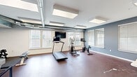 This fitness center in Memphis, TN offers a comprehensive workout environment designed to accommodate a variety of exercise routines. The space is laid out on a single floor and is divided into distinct functional zones. The cardio area features a treadmill, elliptical trainer, and stationary spin bike, each accompanied by individual monitor screens for entertainment during workouts. A wall-mounted flat-screen TV is also positioned for easy viewing across the room. The strength and flexibility zone is equipped with a weight bench, a set of dumbbells, kettlebells, and workout mats, providing ample options for resistance and floor-based training. Wall-mounted racks hold rolled exercise mats and resistance bands, keeping the space organized. A separate open area with a folding table and chairs offers a flexible multipurpose space suitable for small group sessions or cool-downs. The flooring throughout is durable rubber-style material in a deep burgundy tone, well-suited for high-impact use. Exposed silver ventilation ductwork runs along the ceiling, complementing the industrial aesthetic, while large windows dressed with white blinds allow natural light to fill the room. Bright overhead panel lighting ensures consistent illumination throughout the facility.