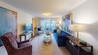 This single-floor residence offers a well-organized layout that balances comfort and style across its key living areas. The living room serves as the heart of the home, featuring plush carpeted flooring, large windows dressed with sheer curtains that invite natural light, and eye-catching large-scale abstract artwork adorning the walls. A tufted navy blue sofa, eclectic accent chairs, and a warm wood-toned media console anchor the space with personality and color.

Adjacent to the living area, a separate dining space is visible, outfitted with a round table and teal upholstered chairs beneath coordinating wall art. The kitchen is practical and well-appointed, featuring warm honey-toned wood cabinetry, white appliances including a range, microwave, and refrigerator, and durable wood-look flooring that adds warmth underfoot.

The primary bedroom provides a serene retreat with a dramatic upholstered headboard, a ceiling fan for comfort, and a window offering a view of lush greenery. A second bedroom mirrors the bold design sensibility with a richly styled navy wingback headboard and coordinating décor. Both bathrooms are clean and functional, featuring warm wood vanities, well-lit mirrors, and tasteful framed artwork. The overall ambiance throughout this residence is vibrant, curated, and welcoming.