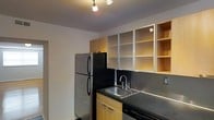 This single-floor space features a thoughtfully designed kitchen that balances functionality with a clean, modern aesthetic. The kitchen is equipped with stainless steel appliances, including a full-sized refrigerator and a dishwasher, making it practical for everyday use. Dark countertops provide a striking contrast against the warm-toned, light wood cabinetry that lines the lower and upper sections of the kitchen. The upper cabinets feature a mix of frosted glass-paneled doors and open shelving, offering both concealed and accessible storage options. A stainless steel sink with a contemporary arc-style faucet is neatly integrated into the countertop. The dark backsplash adds depth and a cohesive design element throughout. Overhead, a track lighting system provides focused, adjustable illumination across the workspace, enhancing both functionality and ambiance. The neutral-toned walls and ceiling contribute to a clean, uncluttered feel. Overall, the kitchen presents a well-organized, contemporary design with durable finishes suited for practical, daily use.