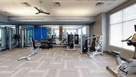 This fitness center offers a comprehensive workout environment designed with both functionality and aesthetics in mind. The space is organized into distinct exercise zones, all unified by neutral-toned herringbone-patterned flooring and a clean, contemporary color palette of gray and blue accents.

The cardio zone features a row of True-brand treadmills, stationary bikes, and elliptical machines, each equipped with individual display screens. Large windows line the walls in this area, allowing abundant natural light to fill the space while roller shades provide adjustable light control. A ceiling fan enhances air circulation throughout the floor.

The strength training area is anchored by a full wall of mirrors that visually expand the space and aid in form monitoring. A well-stocked dumbbell rack, cable machines, and selectorized strength equipment are thoughtfully arranged to allow ease of movement between stations. A dedicated rack holds stability balls and resistance accessories for functional training.

A warm wood-paneled column serves as a striking architectural accent, adding visual contrast to the otherwise contemporary interior. Bright linear lighting fixtures provide consistent illumination throughout. A glass-enclosed room visible near the entry appears to offer a separate training or stretching area, adding further versatility to the facility.