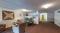 This single-floor space presents a thoughtfully designed interior that balances functionality with a warm, welcoming ambiance. The central lounge area serves as a comfortable gathering point, furnished with upholstered armchairs and a side table, arranged to encourage relaxed conversation or quiet rest. A tall, lush indoor plant adds a natural, refreshing element to the space, complementing the warm-toned, patterned carpet that runs throughout the floor.

Soft, ambient lighting is provided by a combination of flush-mounted ceiling fixtures and a stylish arc floor lamp, casting a gentle glow that enhances the inviting atmosphere. The textured white ceiling and neutral-toned walls contribute to a clean, understated aesthetic throughout.

A partial dividing wall creates a subtle separation between the lounge and the adjacent corridor, which extends further into the facility. This hallway is well-lit and lined with warm wood-toned doors, suggesting access to private rooms or dedicated spaces beyond. Framed artwork along the walls adds a decorative touch, lending a refined character to the overall environment. The layout is open and easy to navigate, making it well-suited for a professional, hospitality, or care-oriented setting.