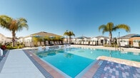 Located at 1201 Cleveland Ave in Wildwood, Florida, this outdoor amenity area showcases a beautifully designed resort-style pool complex that serves as a central gathering space for the community. The expansive swimming pool features a gradual entry point, clearly marked depth indicators, and crystal-clear blue water that reflects the abundant Florida sunshine. Surrounding the pool, a generously sized paver deck is finished in a combination of tile and interlocking brick patterns, adding both visual interest and durability underfoot. A long row of cushioned lounge chairs lines the perimeter of the pool deck, complemented by umbrella-shaded seating clusters that provide relief from the sun. Tall, mature palm trees are strategically placed throughout the area, lending a tropical ambiance and natural shade. The pool area is fully enclosed by a sleek black metal fence for safety and privacy. At one end of the complex sits a well-appointed single-story clubhouse with white clapboard siding, a distinctive cupola, and a covered porch with columns — offering a welcoming space for residents to gather. Lush landscaping with tropical plantings borders the hardscape, tying together the overall aesthetic. The surrounding community features neatly constructed residential homes, all visible from the pool area under the open Florida sky.