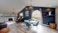 This thoughtfully designed single-floor space offers an impressive open layout defined by soaring vaulted ceilings and an array of architectural details that create a welcoming, upscale atmosphere. Light hardwood flooring runs throughout, unifying the various functional zones seamlessly.

The main gathering area commands attention with a bold navy blue accent wall that rises to meet the pitched ceiling, flanked by large arched windows dressed in patterned drapes. A leather sectional sofa anchors the space beneath an orb-style pendant chandelier, while a stacked-stone fireplace with a wood mantel serves as a striking focal point along an adjacent wall, complemented by abundant natural light from multiple windows.

A defined dining area flows naturally from the lounge, featuring a round pedestal table surrounded by teal velvet chairs beneath a matching chandelier. Beyond, a reception zone centers on a substantial built-in counter beneath a generous skylight, flooding the area with natural light. French doors lead to an adjoining room, adding further functionality.

The welcoming entryway includes a console table, seating, and signage indicating tours are available, suggesting this space serves as a community or leasing facility. Throughout, the combination of rich color, natural textures, and carefully considered lighting creates a refined yet comfortable ambiance.