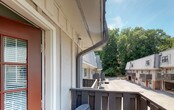 This single-floor unit is part of a charming multi-unit residential community characterized by a distinctive combination of vertical board-and-batten siding, brick base accents, and standing-seam metal roofing. Dormer windows add architectural interest to the roofline, enhancing the overall aesthetic of the complex. A bold red entry door with divided-light glass panels provides a welcoming focal point and leads to the unit's private outdoor deck. The deck is a notable feature, finished with wood decking and railing, and furnished with seating that invites relaxation. From the deck, residents enjoy open views of lush, mature tree canopies that provide a natural, tranquil backdrop. The surrounding landscape includes tiered stone retaining walls and fenced yard sections, contributing to a well-organized outdoor environment. The community features a shared paved courtyard area, and a basketball hoop is visible, offering recreational opportunity within the complex. Additional units in the community mirror the same architectural style, creating a cohesive and visually unified appearance. The setting feels tucked away and peaceful, with abundant greenery framing the property on multiple sides, making it an inviting residential environment with both functional outdoor space and natural surroundings.