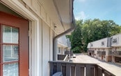 This single-floor unit is part of a charming multi-unit residential community characterized by a distinctive combination of vertical board-and-batten siding, brick base accents, and standing-seam metal roofing. Dormer windows add architectural interest to the roofline, enhancing the overall aesthetic of the complex. A bold red entry door with divided-light glass panels provides a welcoming focal point and leads to the unit's private outdoor deck. The deck is a notable feature, finished with wood decking and railing, and furnished with seating that invites relaxation. From the deck, residents enjoy open views of lush, mature tree canopies that provide a natural, tranquil backdrop. The surrounding landscape includes tiered stone retaining walls and fenced yard sections, contributing to a well-organized outdoor environment. The community features a shared paved courtyard area, and a basketball hoop is visible, offering recreational opportunity within the complex. Additional units in the community mirror the same architectural style, creating a cohesive and visually unified appearance. The setting feels tucked away and peaceful, with abundant greenery framing the property on multiple sides, making it an inviting residential environment with both functional outdoor space and natural surroundings.