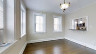 This single-floor space exudes a timeless, understated elegance through its carefully considered architectural details and neutral palette. The main living area showcases rich, dark hardwood flooring that anchors the room with warmth and character, complemented by crisp white baseboards and classic crown molding that frame the space with refined definition. Soft beige walls create a welcoming, neutral backdrop that adapts easily to a variety of furnishing styles. Natural light pours generously into the room through multiple white-framed double-hung windows dressed with slatted blinds, offering both brightness and privacy as needed. A decorative pendant light fixture hangs from the ceiling, adding an ornate focal point overhead and lending the space a touch of traditional charm. The ceiling itself features a subtle textured finish, adding visual interest above. A pass-through opening along one wall hints at an adjoining room, suggesting a practical and connected layout that encourages an easy flow between spaces. Overall, the space presents a clean, move-in-ready environment with classic bones and a calm, inviting ambiance suited to a range of uses.