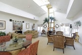 This single-story space is designed with an open, airy layout that emphasizes generous volume and natural light. Soaring ceilings rise dramatically throughout the main living areas, creating a grand sense of scale. Large windows dressed with neutral Roman shades line the walls, framing outdoor views of surrounding greenery while flooding the interiors with warm, diffused light.

The main gathering area features a pair of plush, earth-toned sofas arranged symmetrically around a decorative black cabinet, flanked by framed artwork in gilded frames and a hanging tapestry that serves as a focal centerpiece. A patterned area rug anchors the seating arrangement and adds warmth to the neutral carpeted flooring found throughout.

An adjacent lounge area showcases a wicker dining set with a glass-topped table, complemented by tall decorative gold vases and wall-mounted floral accents. The space flows naturally into a striking sunlit sitting area featuring towering indoor palm trees that reach toward the soaring ceiling, creating a dramatic tropical focal point. Elegant accent chairs, an ornate area rug, and decorative shelving complete this area.

A chandelier adds a refined touch overhead, while recessed lighting provides additional illumination. A glass door leads to an outdoor patio, further enhancing the connection between the interior and exterior environments. Overall, the space exudes a warm, eclectic elegance.