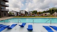 Located at 2508 April Liu Ln. in Charlotte, NC, this single-floor community amenity space offers a well-rounded set of features designed for comfort and convenience. The exterior centerpiece is a large, inviting outdoor swimming pool with a broad, flat pool deck lined with blue lounge chairs and cozy pod-style seating areas set against a manicured lawn. The pool area provides open views of the surrounding multi-story residential buildings and mature tree line, creating a pleasant outdoor retreat.

Inside, the space is defined by clean white walls, high ceilings, and patterned carpet flooring that runs throughout the interior corridors. The hallways are wide and well-lit, with recessed and flush-mounted ceiling fixtures providing consistent illumination. Accessibility is a clear priority, with ADA-compliant signage and features visible near the restroom entrances. Glass-panel doors at the end of the main corridor allow natural light to filter in while offering direct sightlines to the outdoor pool area, creating a seamless visual connection between the interior and exterior spaces. The gallery-style main room features artwork displayed on crisp white walls, adding a touch of character to the functional layout. Overall, the space presents a clean, modern, and community-focused environment.