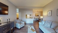 This single-floor residence in Louisville, Kentucky offers a well-designed, open-concept layout that connects the living, dining, and kitchen areas seamlessly. The living room features rich hardwood-style flooring, neutral walls, and comfortable seating, creating a welcoming atmosphere. It flows naturally into the dining area, which is anchored by an elegant chandelier overhead and opens directly to the kitchen. The galley-style kitchen is outfitted with dark espresso cabinetry, granite-look countertops, a stainless sink, and white appliances, providing ample storage and functional workspace. The primary bedroom is carpeted for warmth and comfort, featuring a walk-in closet and natural light from a window with sheer curtains. A secondary bedroom also offers carpeted flooring and closet access, while an additional room serves as a dedicated office or flex space. Two full bathrooms are finished with wood-look flooring, dark vanity cabinetry, granite countertops, and tub-shower combinations, delivering a cohesive and polished aesthetic. Throughout the residence, neutral tones and consistent finishes create a calm, unified ambiance suited for everyday living in the heart of Louisville.